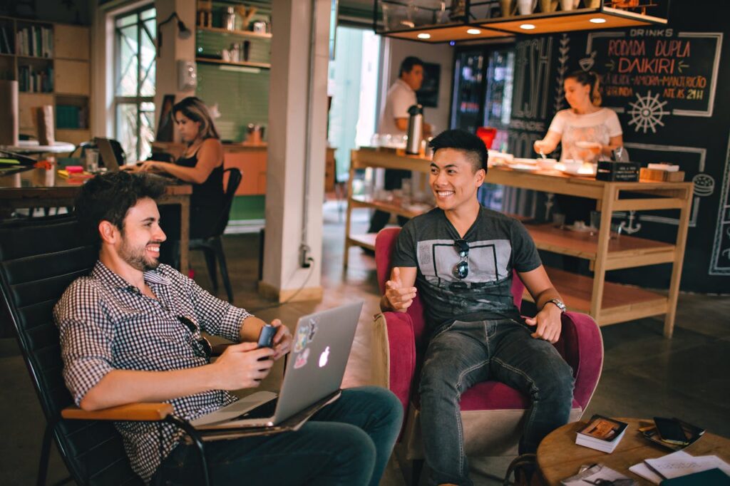 A imagem mostra um ambiente descontraído com pessoas conversando e trabalhando em seus notebooks, representando as vantagens de buscar as comunidades de nômades digitais enquanto estiver viajando e trabalhando.